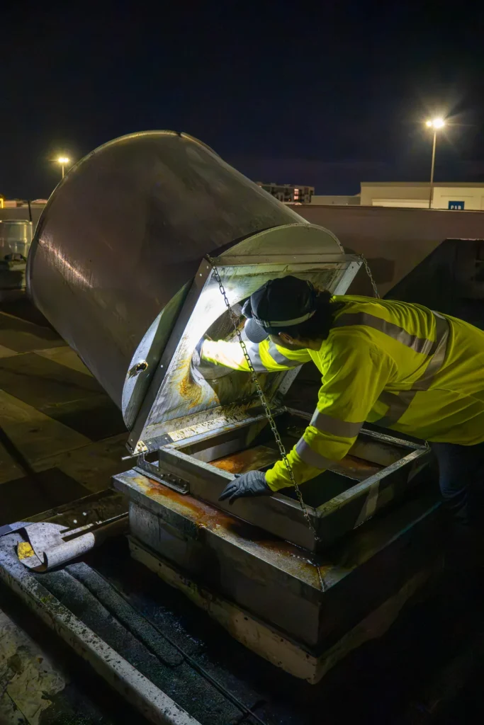 Technician with a headlamp inspecting grease buildup inside a hinged-open rooftop exhaust fan before cleaning.